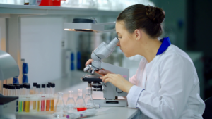 Female scientist in a lab coat looking through a microscope, surrounded by test tubes and glass beakers, illustrating clinical research into inherited retinal diseases.